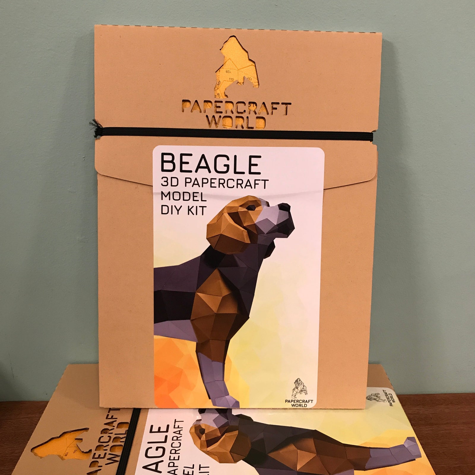Packaging for the Paper Craft World Beagle Kit. It is kraft paper with the papercraft world logo laser cut into the packaging. It has a sticker on the front with a picture of the completed beagle craft kit.