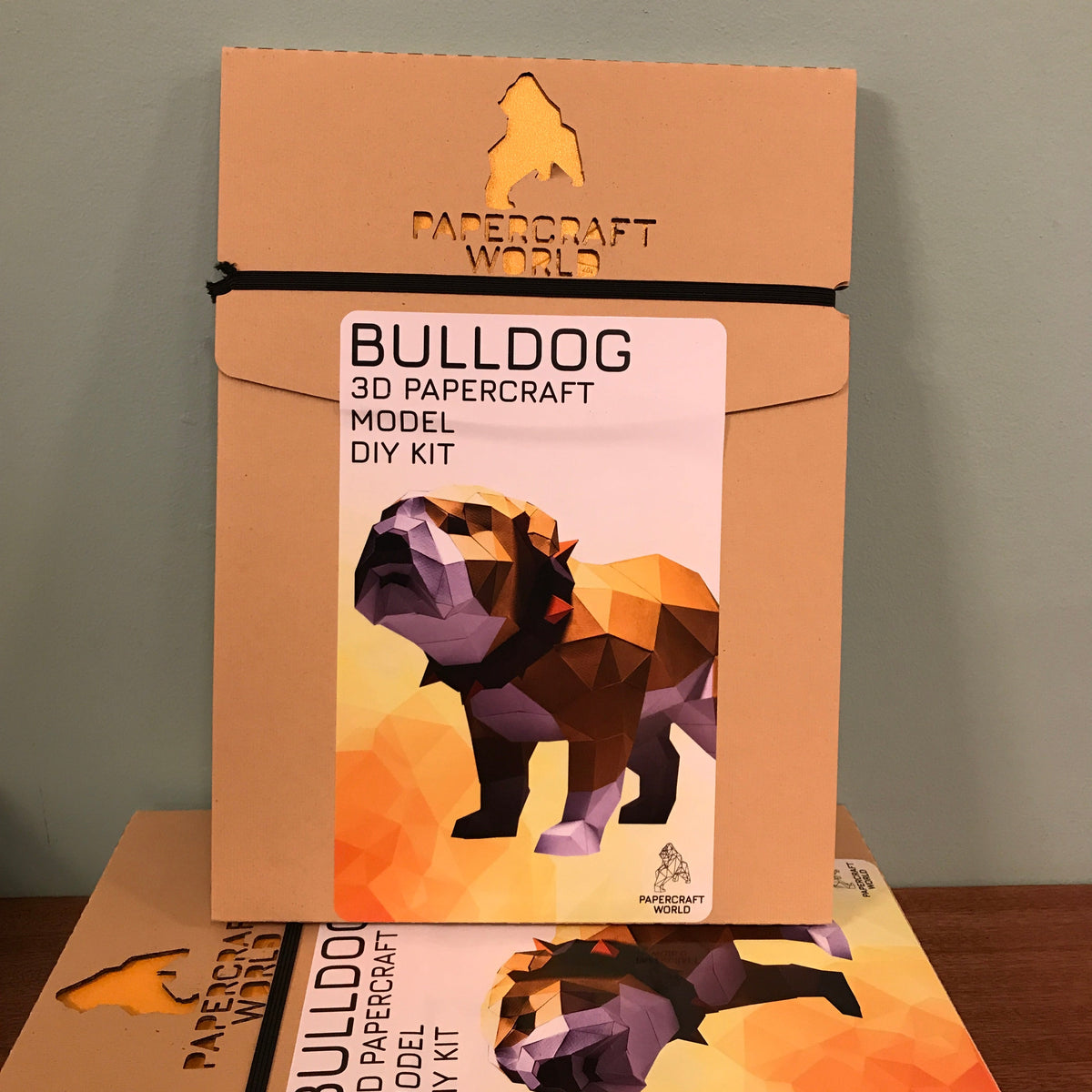 The packaging for the bulldog paper craft kit. It is kraft colored with the papercraft world logo laser cut into the top of the package. There is a photo of a completed example of a bulldog paper craft kit against a white background.