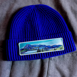 Blue beanie with a mountain landscape patch sitting on a gray jacket. 