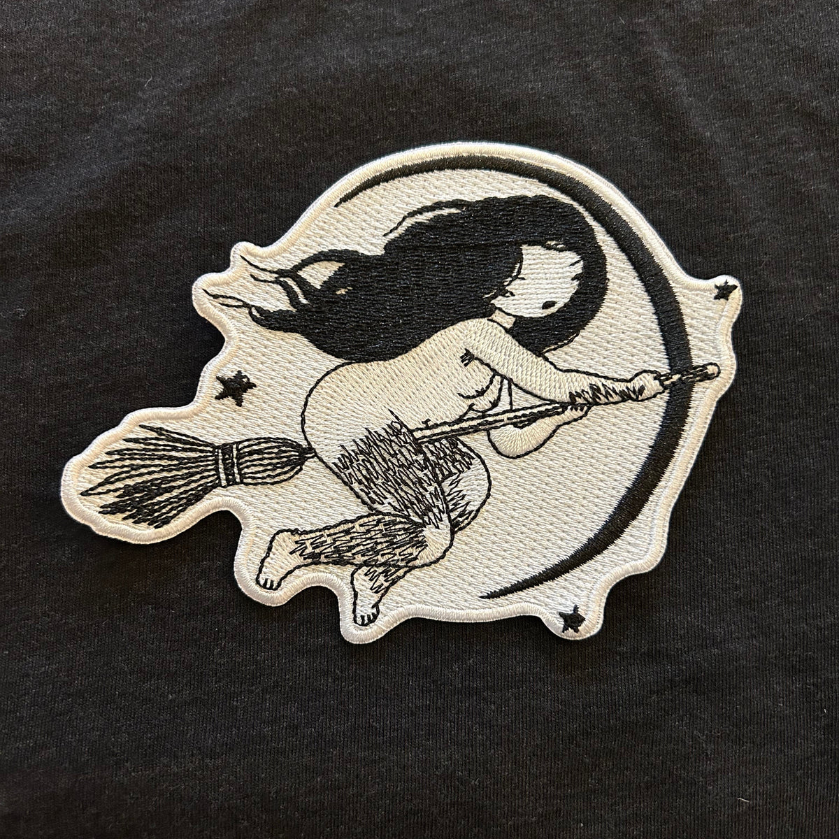 Embroidered design of a witch with hairy legs on a broomstick on a black fabric background. 