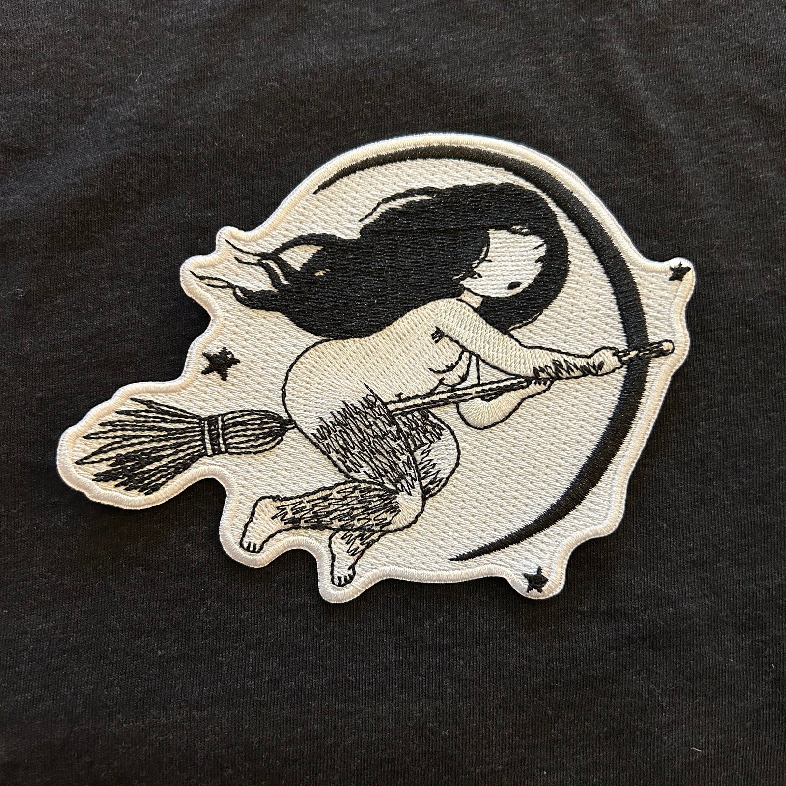 Embroidered design of a witch with hairy legs on a broomstick on a black fabric background. 