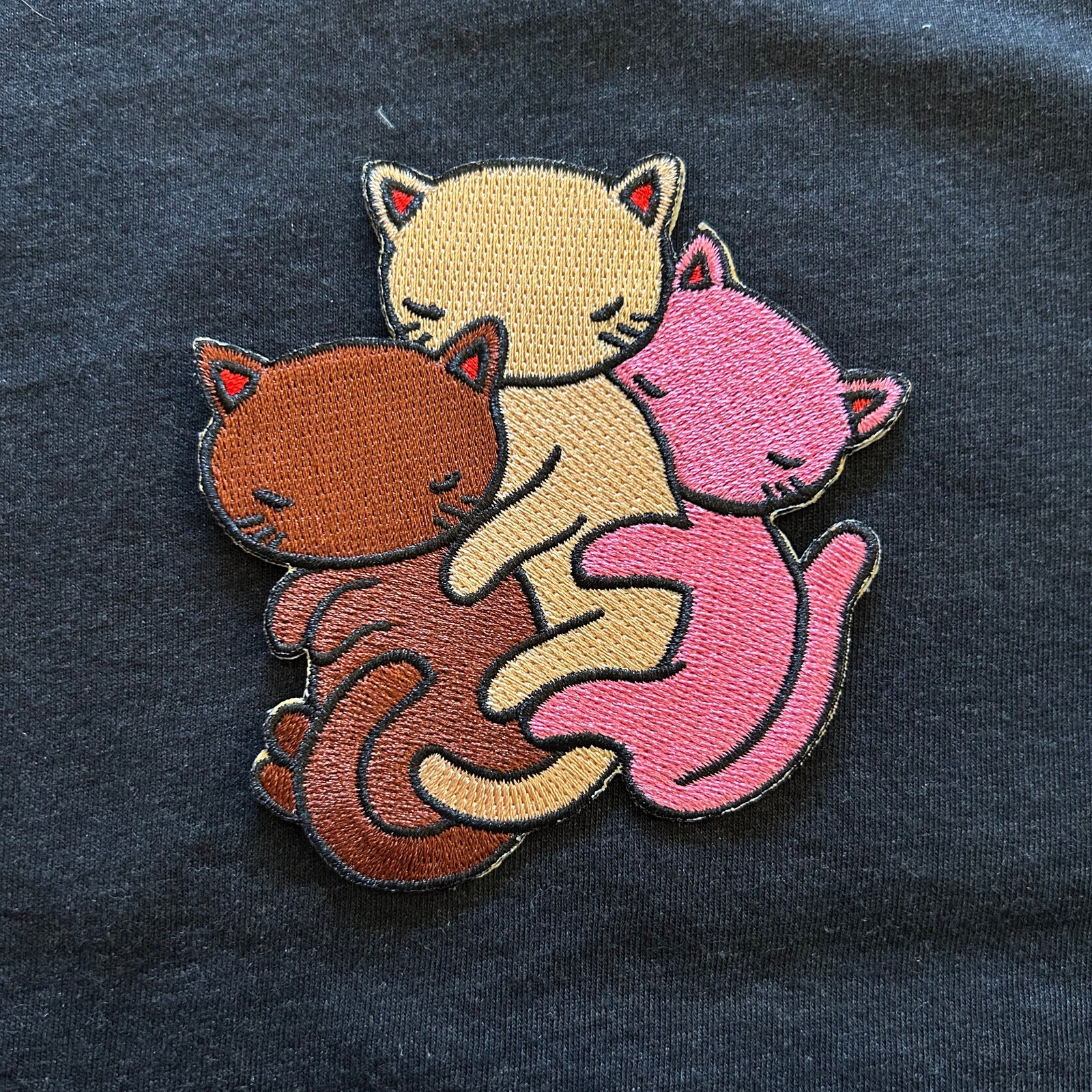 Embroidered patch of three cats in a huddle on a dark fabric background. 