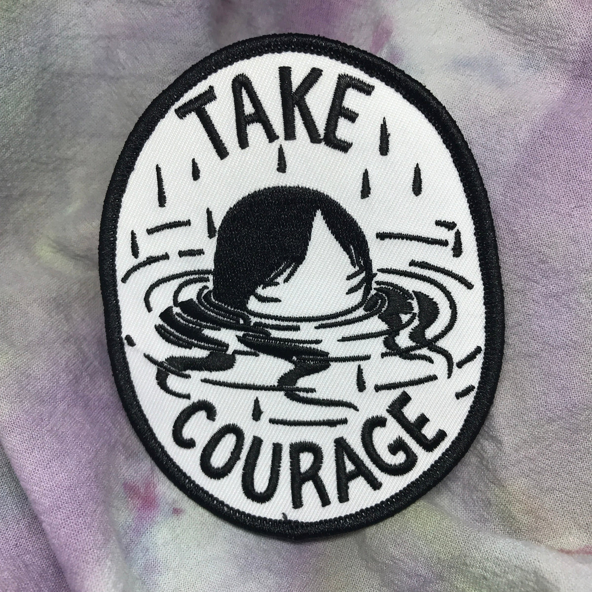 White patch with black stitching. It says Take Courage and has a woman up to her nose in water with raindrops coming down in the background. 