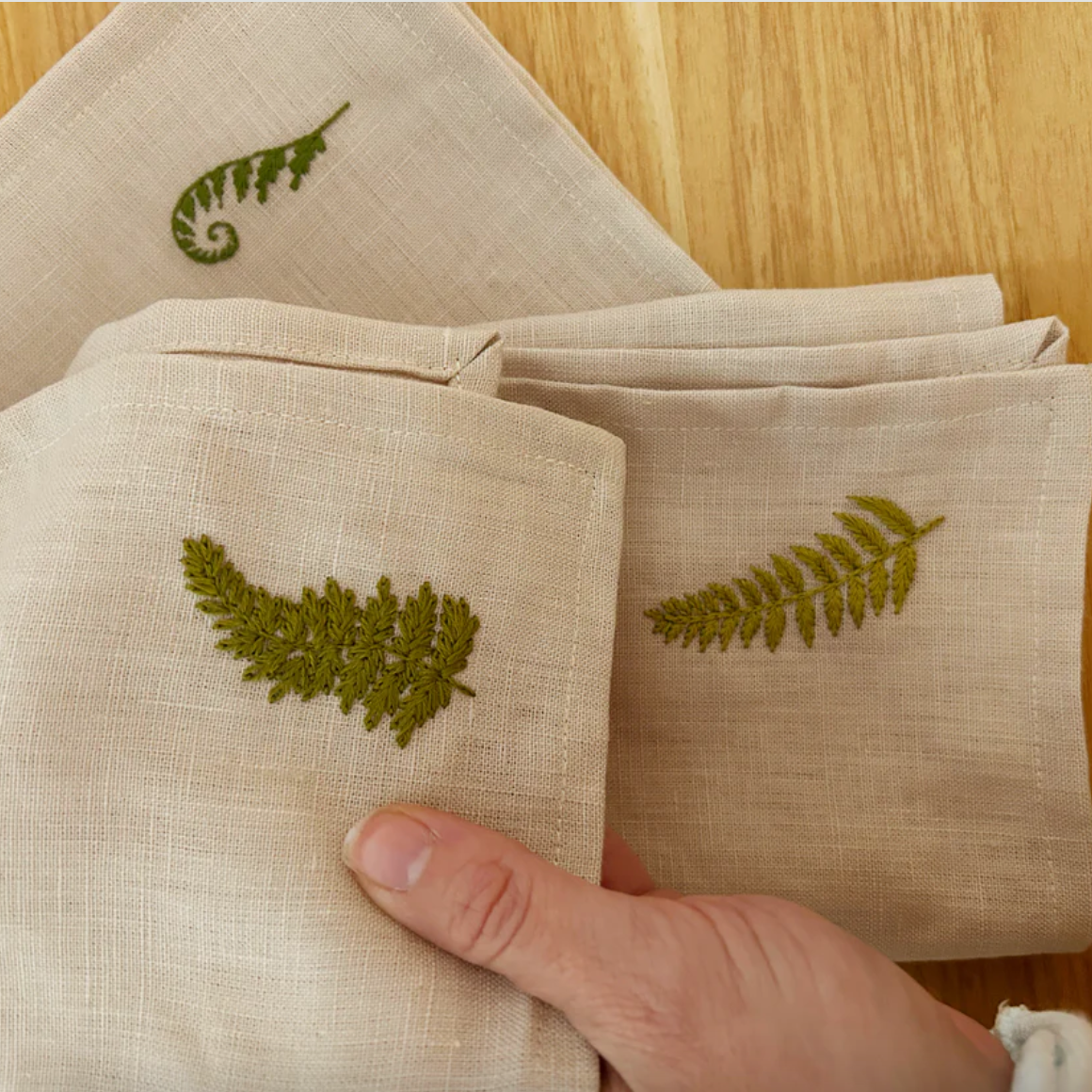 Beige napkins with green leaf embroidery on a wooden surface. 