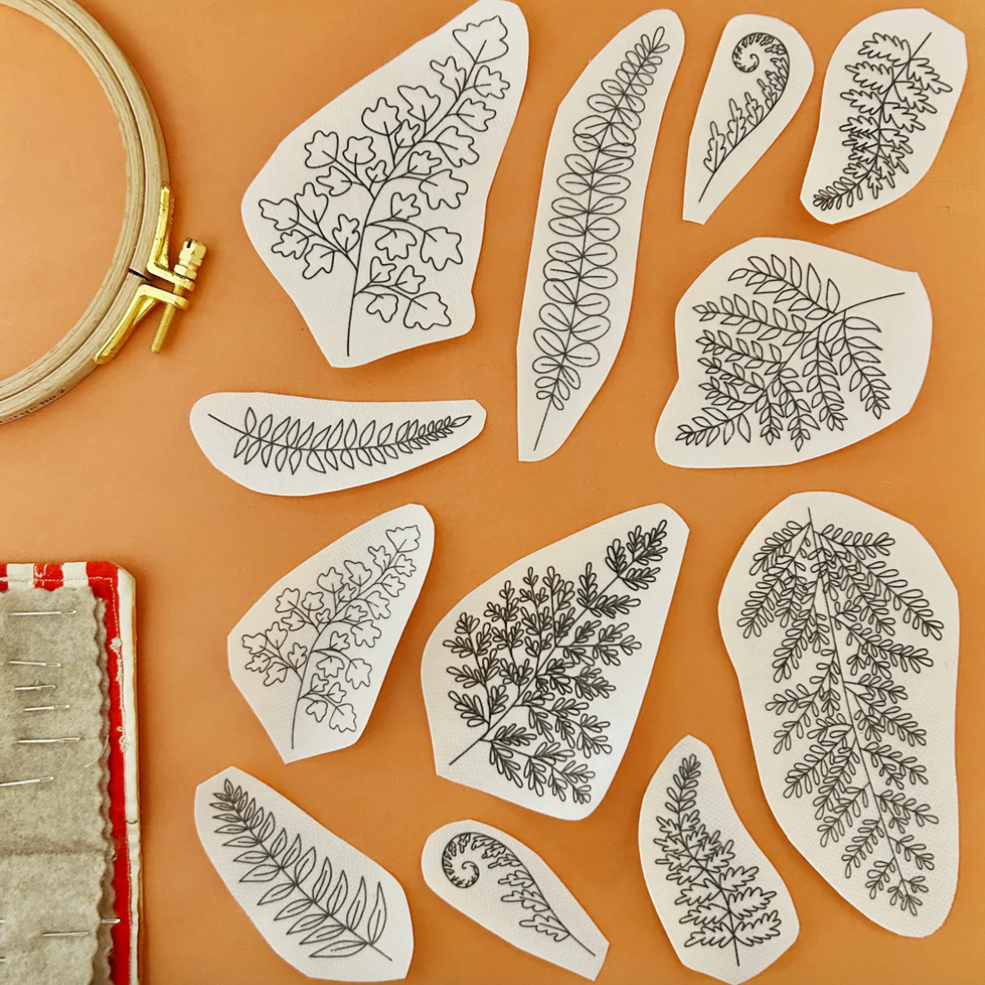 Set of embroidered leaf patterns on fabric with an embroidery hoop and fabric roll on a brown background. 