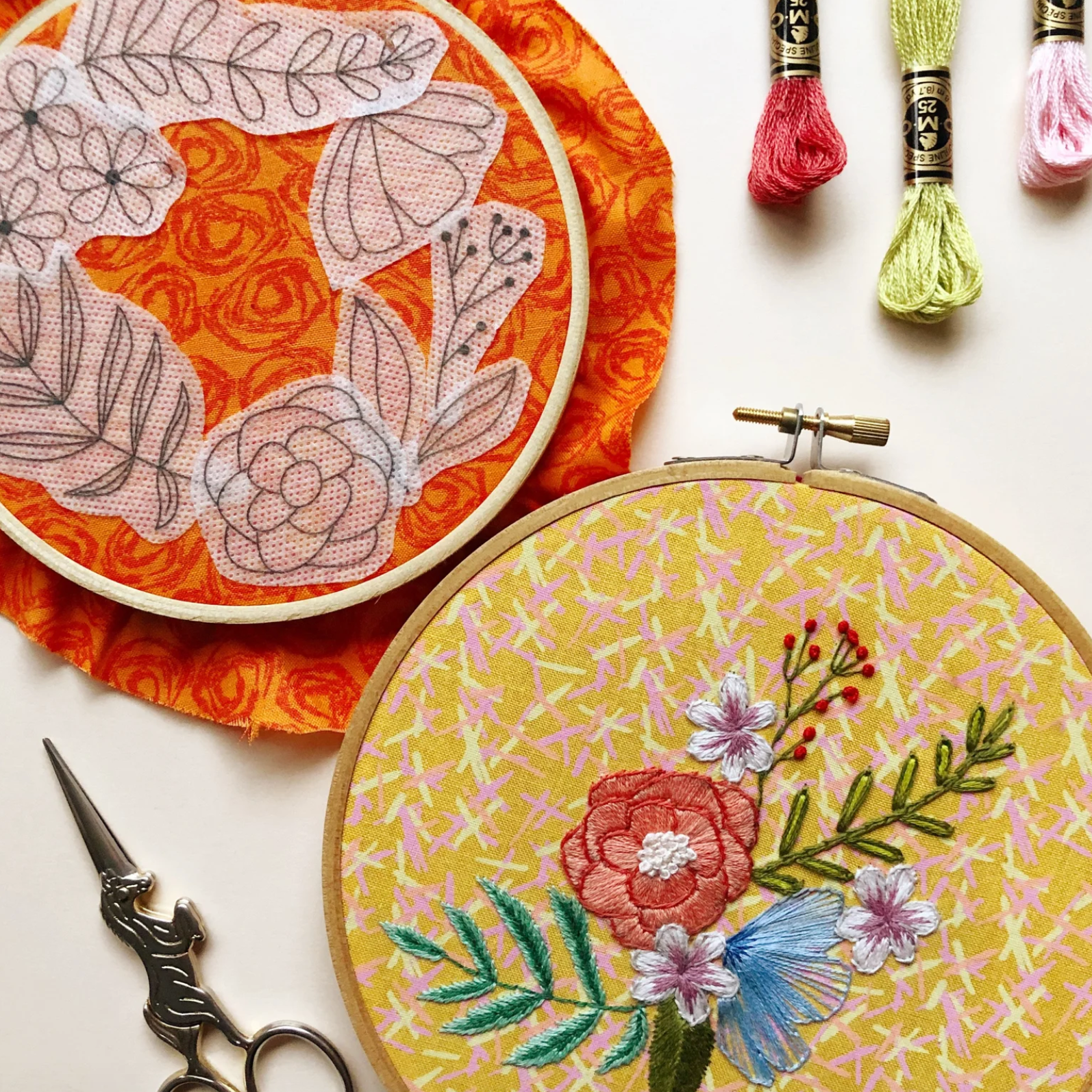 Two embroidery hoops with floral patterns, a pair of scissors, and thread spools on a white background.
