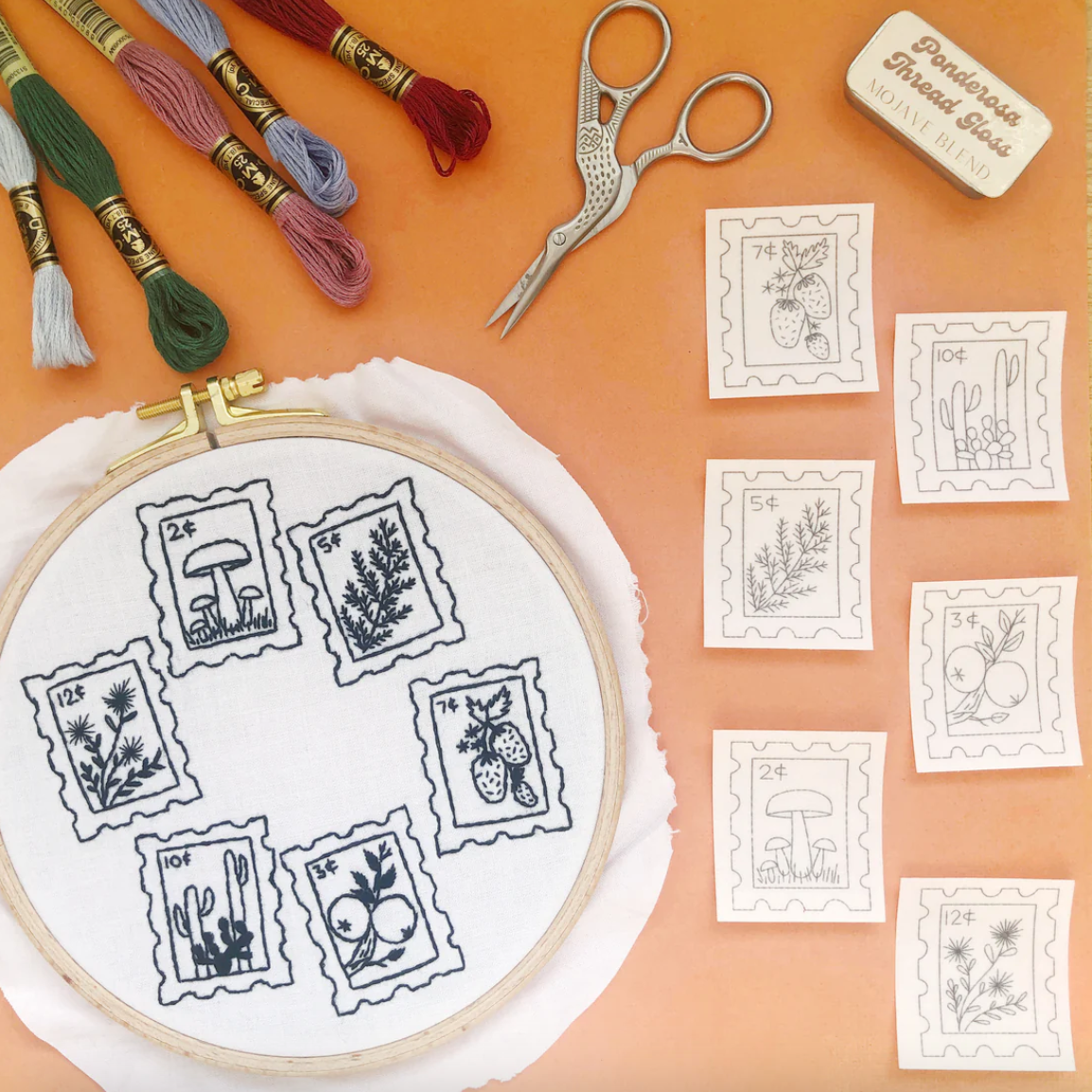 Embroidery hoop with stitched designs, embroidery threads, scissors, and pattern sheets on a peach background. 