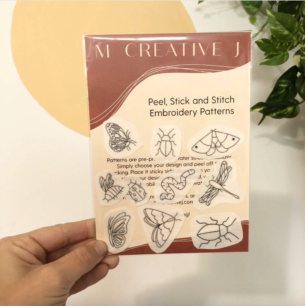 Pack of peel, stick, and stitch embroidery patterns held by a hand with a plant in the background.