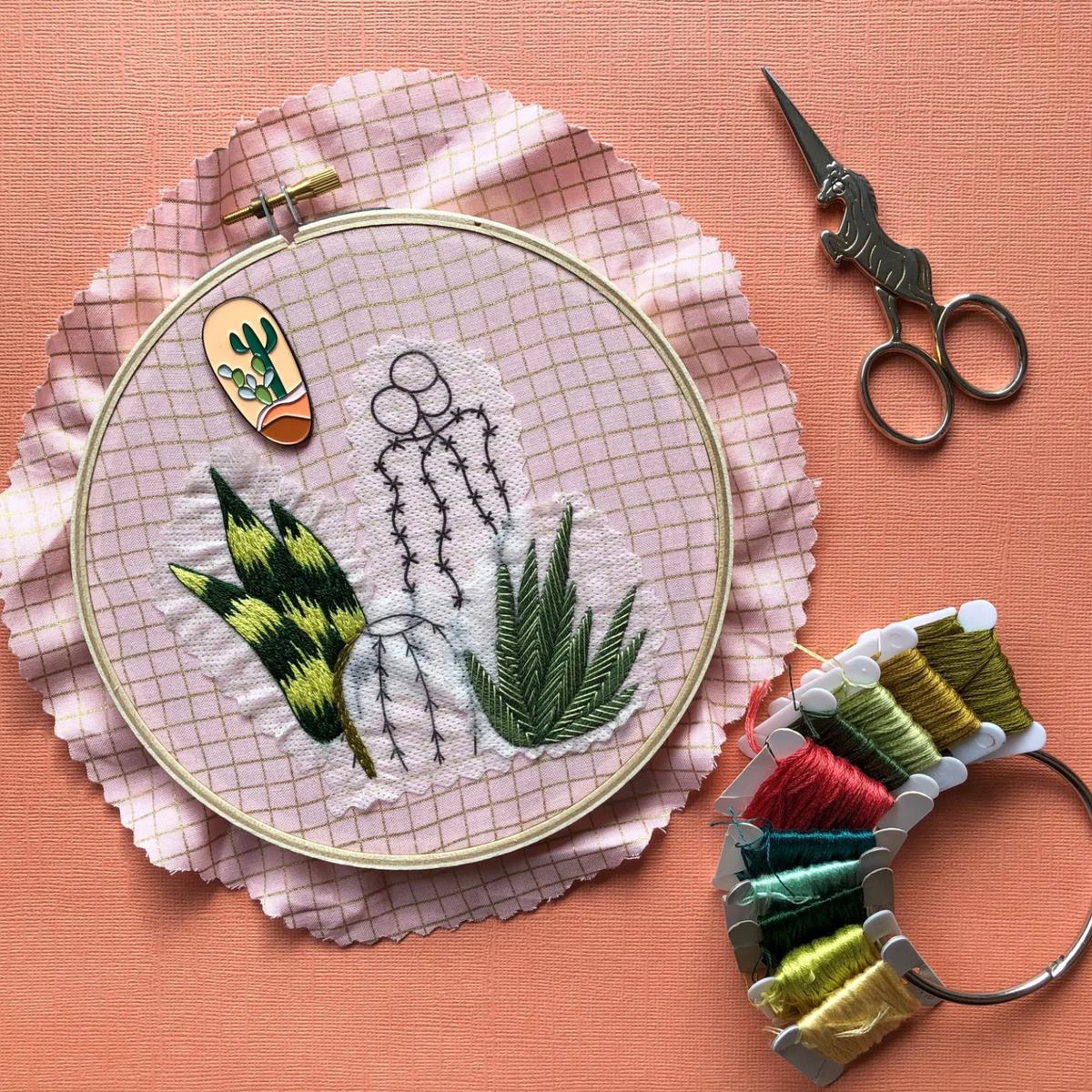 Embroidery hoop with plant designs, scissors, and thread on a pink background