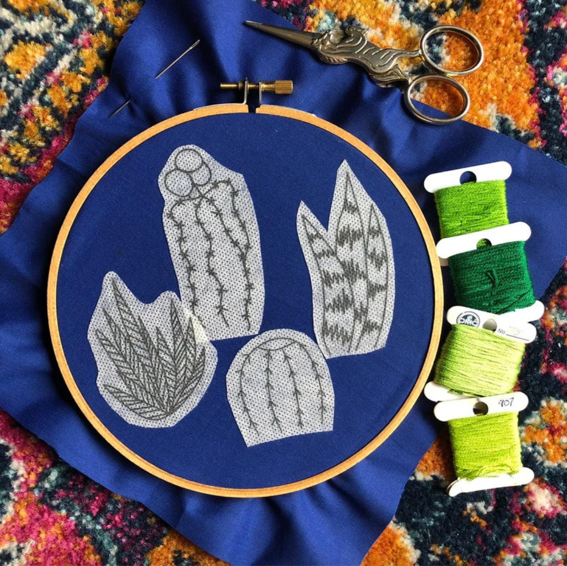 Embroidery hoop with embroidered cacti design on a blue fabric background, surrounded by embroidery supplies.