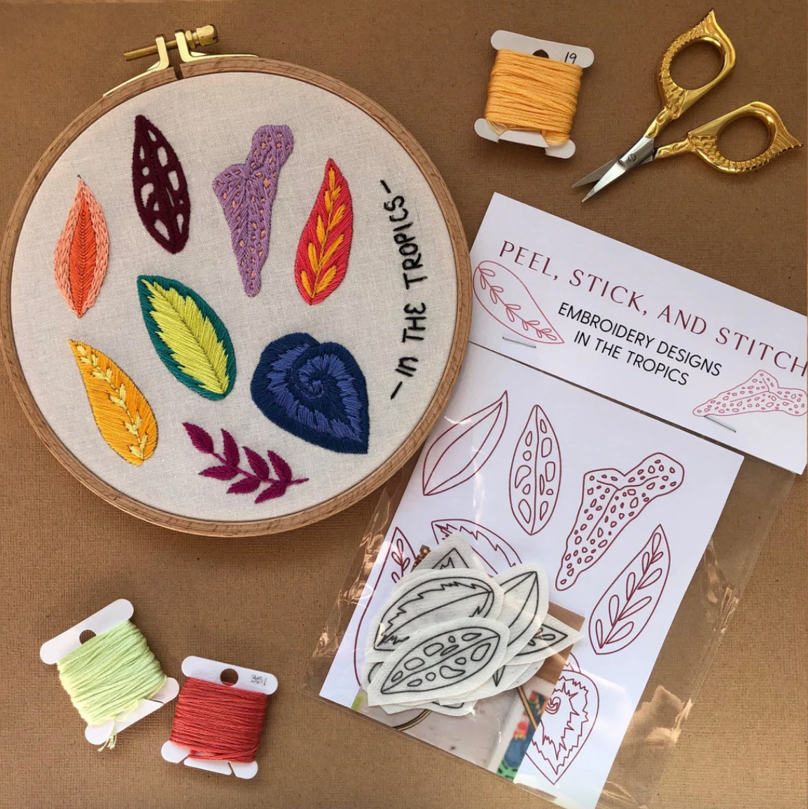 Embroidery hoop with colorful leaves, embroidery threads, scissors, and pattern sheets on a brown background.