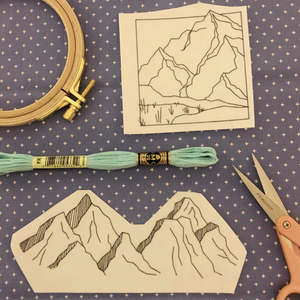 Embroidery hoop, thread, scissors, and mountain-themed fabric pieces on a patterned background. 
