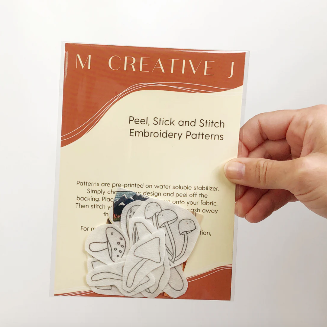 Hand holding a 'M Creative J' embroidery pattern packet with peel, stick, and stitch designs.