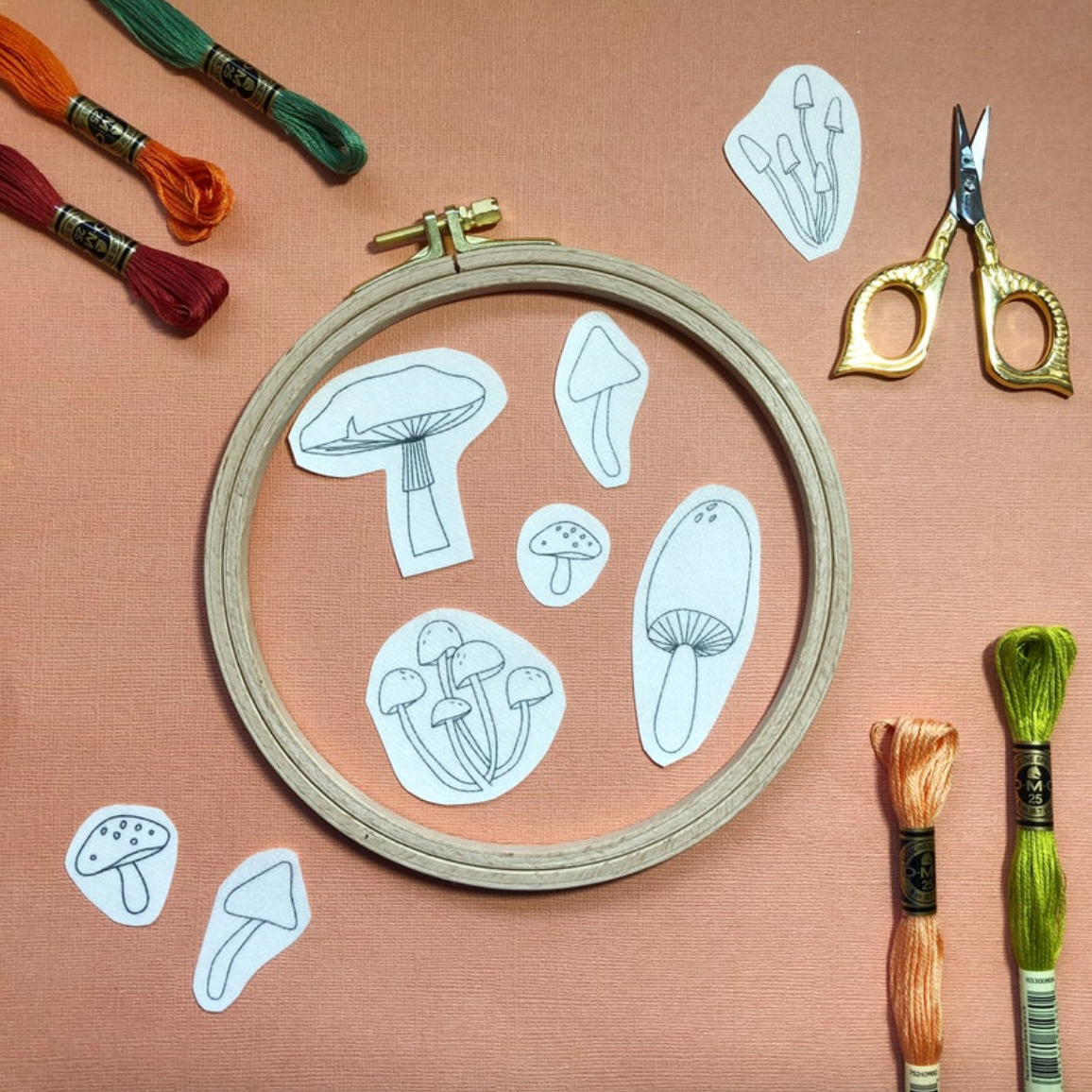 Embroidery hoop with mushroom patterns, embroidery threads, scissors, and pins on a pink background. 