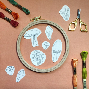 Embroidery hoop with mushroom patterns, embroidery threads, scissors, and pins on a pink background. 