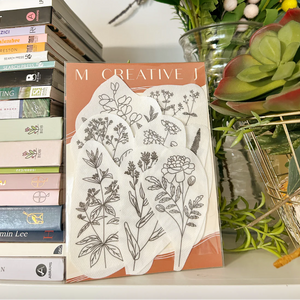 Embroidery supplies with natural dye plant  design on a surface with books and plants in the background. 