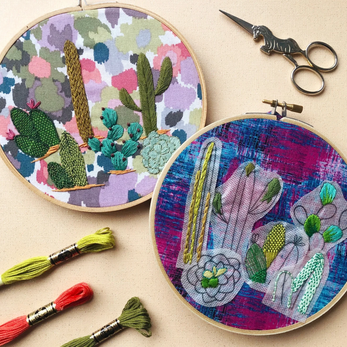 Two embroidery hoops with colorful cactus designs on a beige surface, accompanied by embroidery threads and scissors.