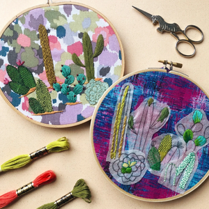 Two embroidery hoops with colorful cactus designs on a beige surface, accompanied by embroidery threads and scissors.