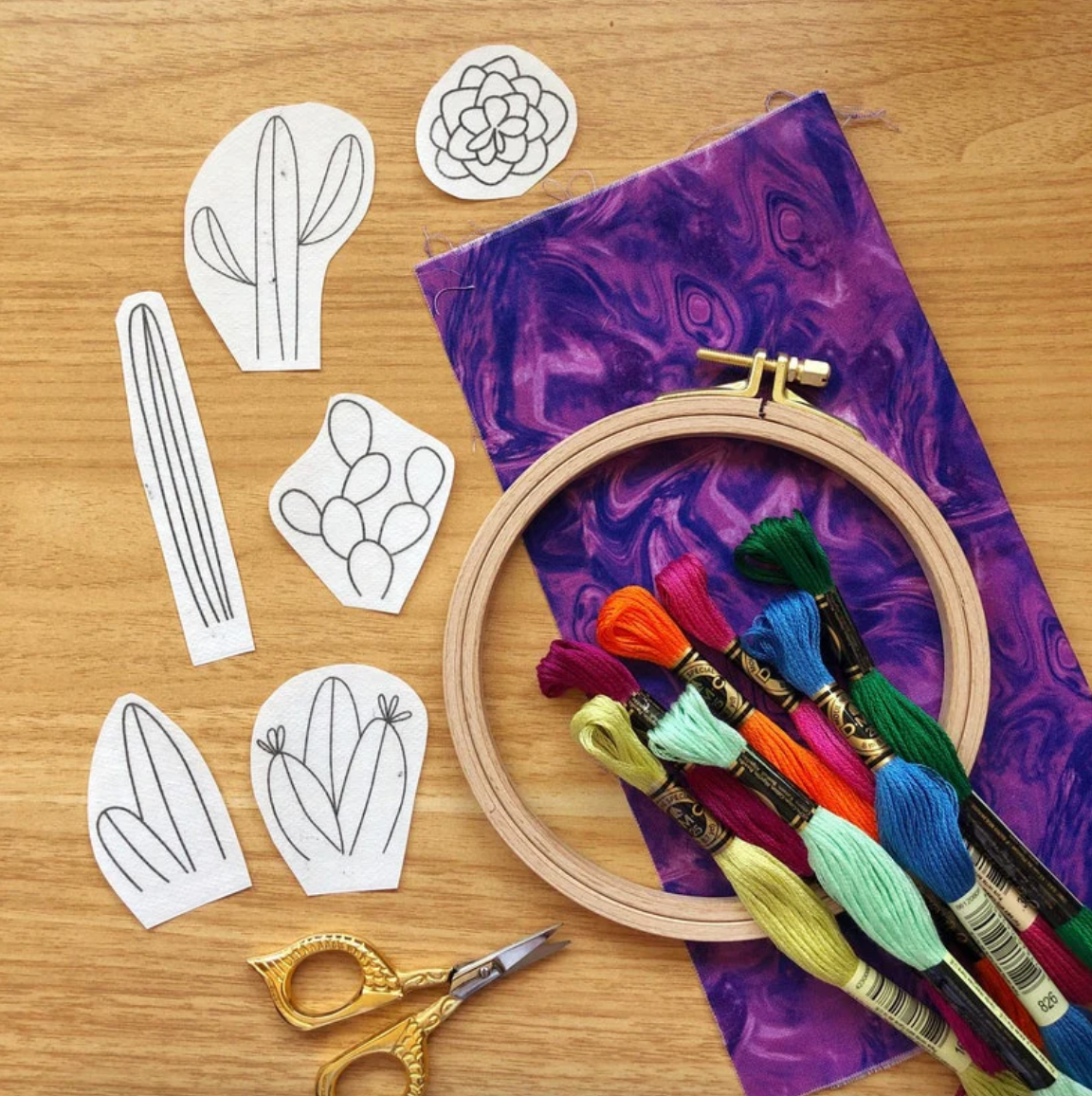 Embroidery hoop with colorful threads, scissors, and cactus-shaped templates on a wooden surface. 