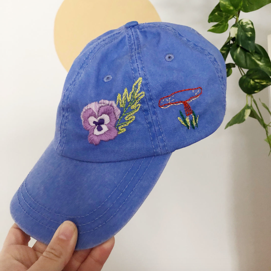 Blue cap with embroidered designs held by a hand against a neutral background. 