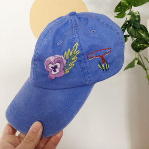 Blue cap with embroidered designs held by a hand against a neutral background. 