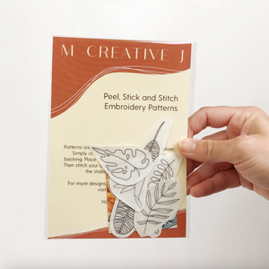 Hand holding a 'M Creative J' embroidery patterns packet with tropical plant designs.