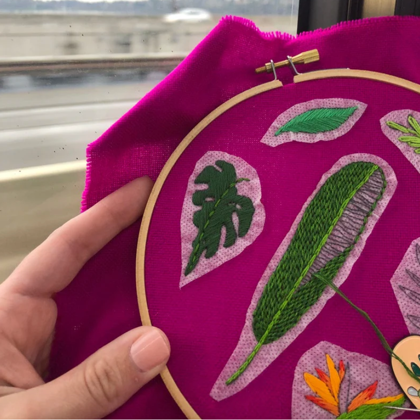Embroidery hoop with green leaves on a purple fabric, held by a hand.