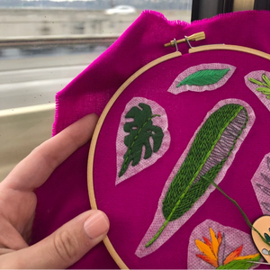 Embroidery hoop with green leaves on a purple fabric, held by a hand.