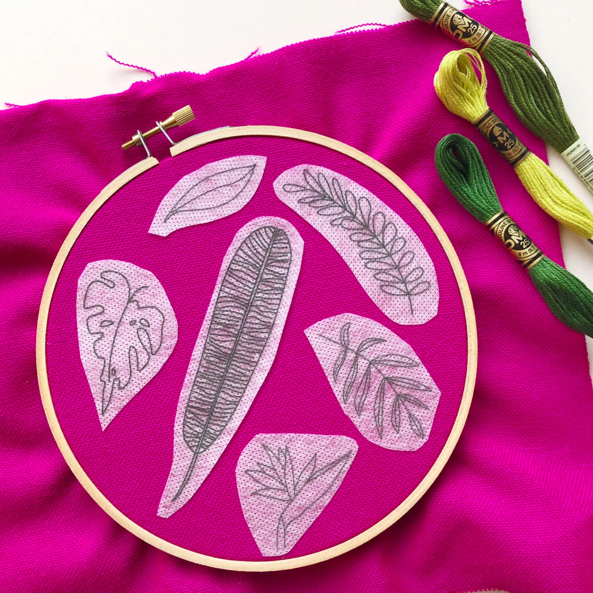 Embroidery hoop with leaf patterns on pink fabric, with embroidery threads on a white background. 