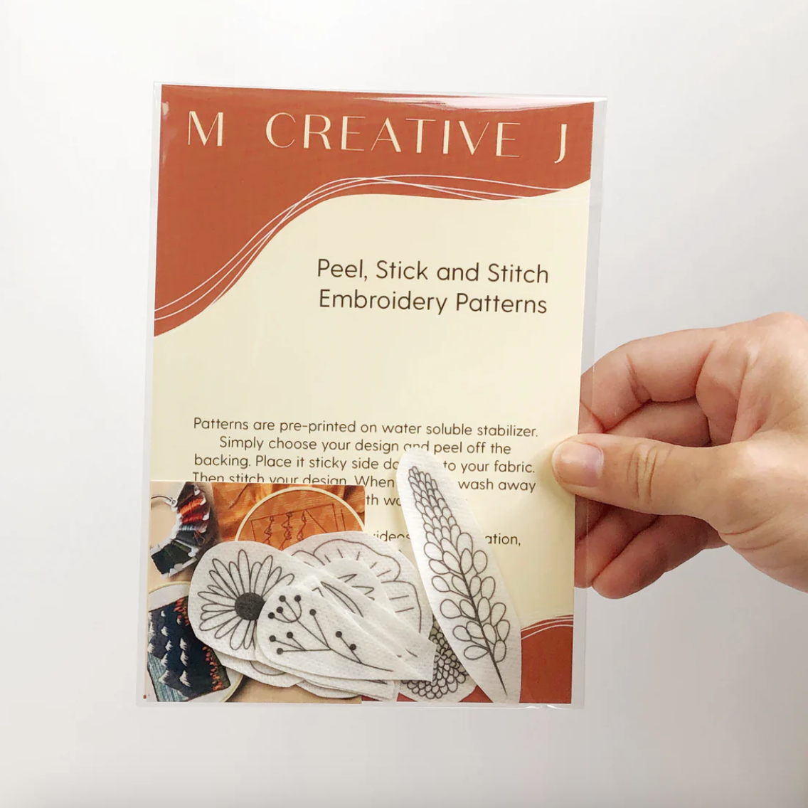 Hand holding a package of 'M Creative J Peel, Stick and Stitch Embroidery Patterns' with wildflower embroidery designs on the cover.