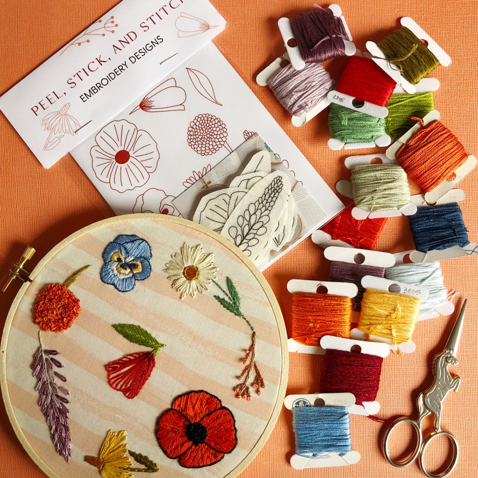 Embroidery hoop with floral design, threads, and pattern book on an orange background. 
