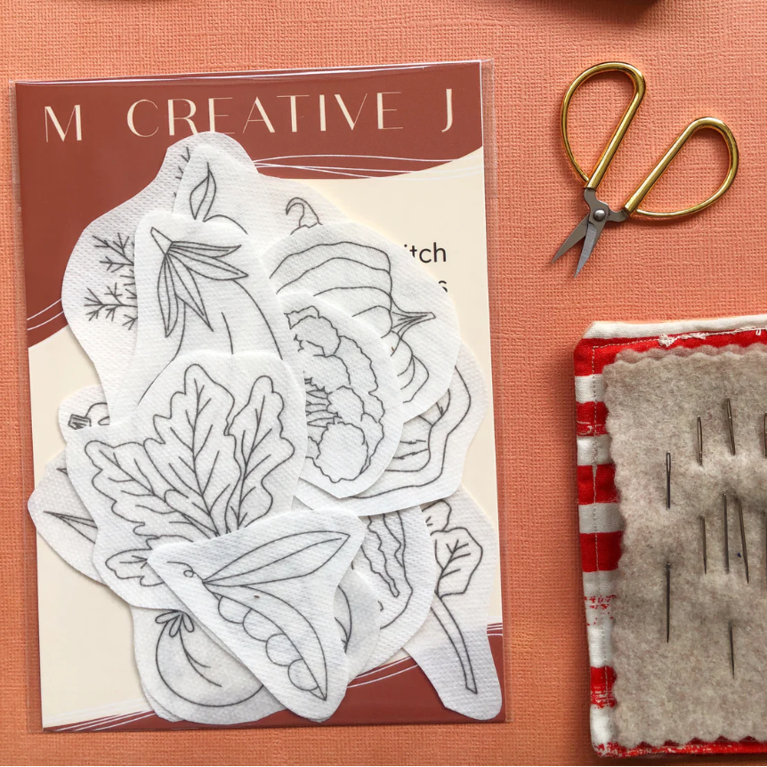 Packaging for vegetable embroidery transfer designs on a pink card, gold scissors, and a red and white patterned block on an orange background.