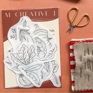 Packaging for vegetable embroidery transfer designs on a pink card, gold scissors, and a red and white patterned block on an orange background.