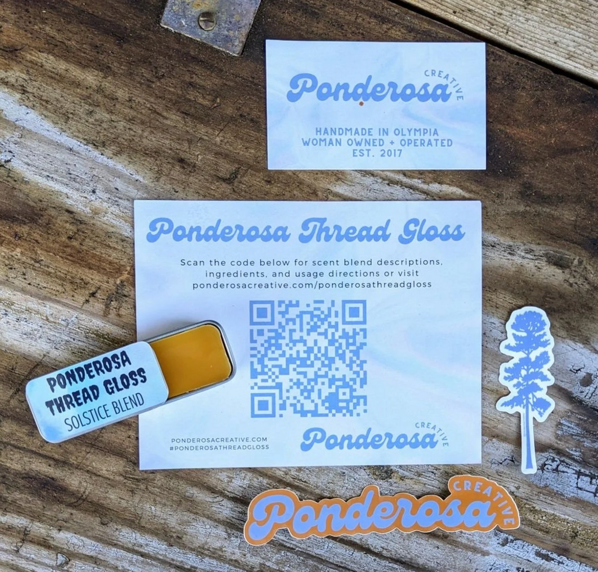 A tin of Ponderosa Thread Gloss against a background with a QR code where you can scan to see the description of the scent and ingredients. There is also a card that says "Ponderosa Creative: Handmade in Olympia. Women Owned + Operated"