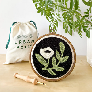 A completed punch needle kit with a white flower on a black background is standing on a table. A cloth bag that says "The Urban Acres" is in the background. A wooden punch needle sits to the side. 