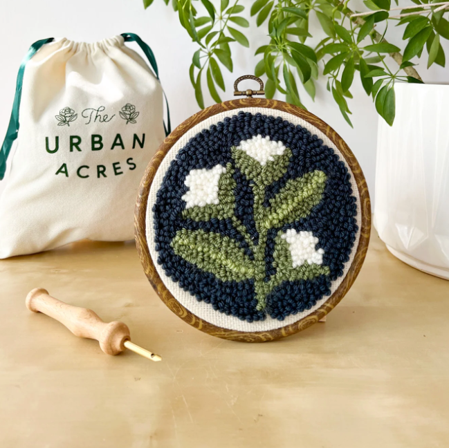 A completed punch needle kit with a white flower on a navy blue background is standing on a table. A cloth bag that says "The Urban Acres" is in the background. A wooden punch needle sits to the side. 