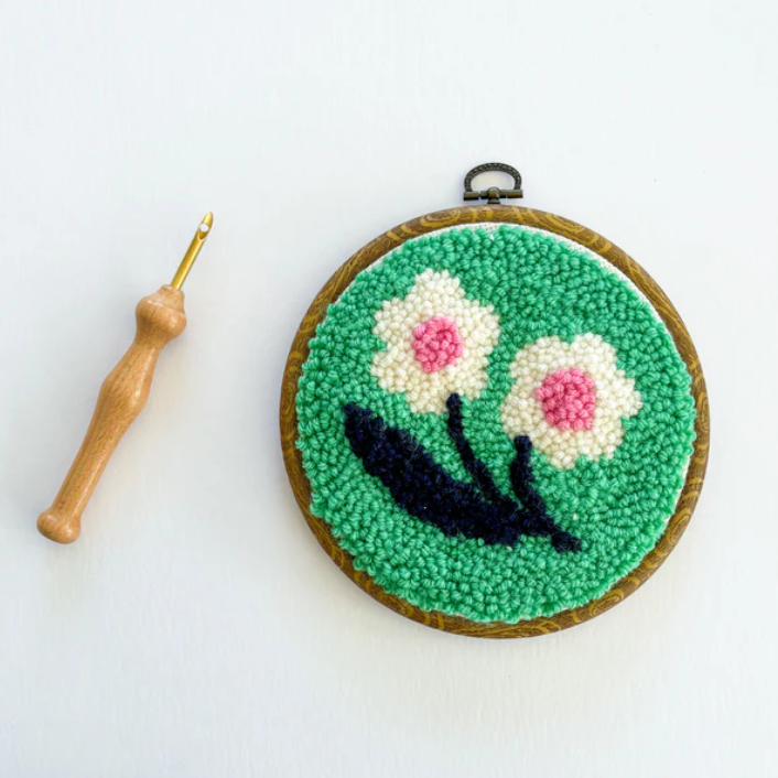 A completed punch needle kit with a white and pink flower and a spring green background is laying on a table. A wooden punch needle sits to the side. 