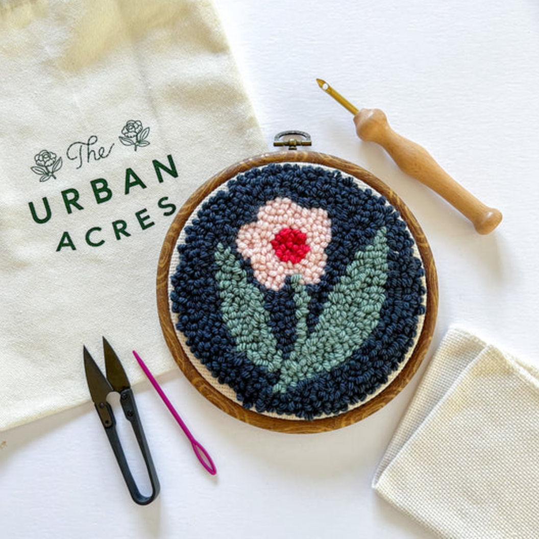 A completed punch needle kit with a pink flower on a navy blue background is laying  on a table. A cloth bag that says "The Urban Acres" is in the background. A wooden punch needle, snips and a yarn needle sit to the side. 
