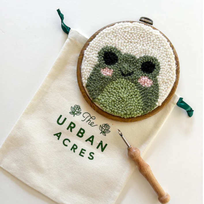 A completed punch needle kit with a frog is laying on a table. A cloth bag that says "The Urban Acres" is in the background. A wooden punch needle sits to the side. 