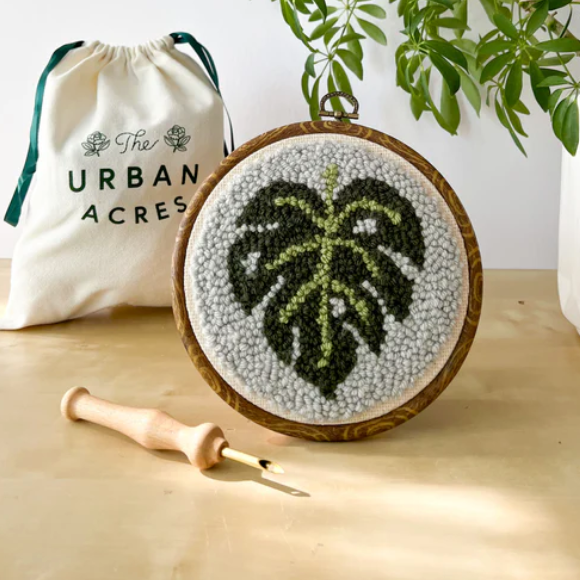 A completed punch needle kit with a monstera leaf is standing on a table. A cloth bag that says "The Urban Acres" is in the background. A wooden punch needle sits to the side. 