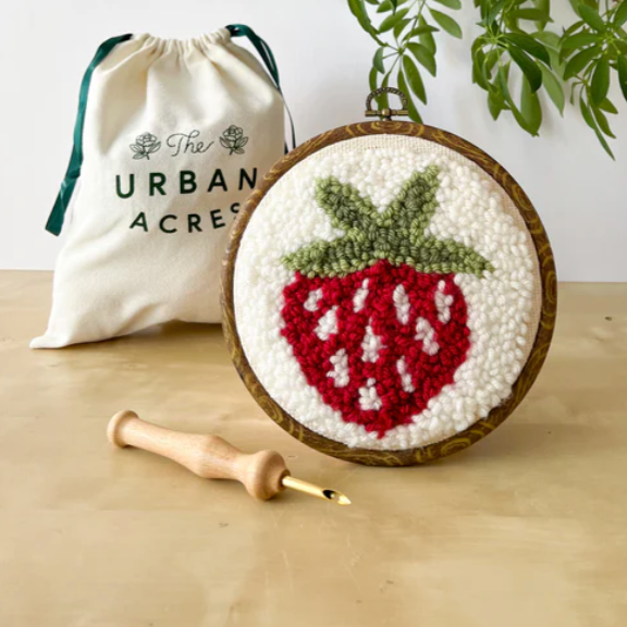 A completed punch needle kit with a juicy strawberry is standing on a table. A cloth bag that says "The Urban Acres" is in the background. A wooden punch needle sits to the side. 