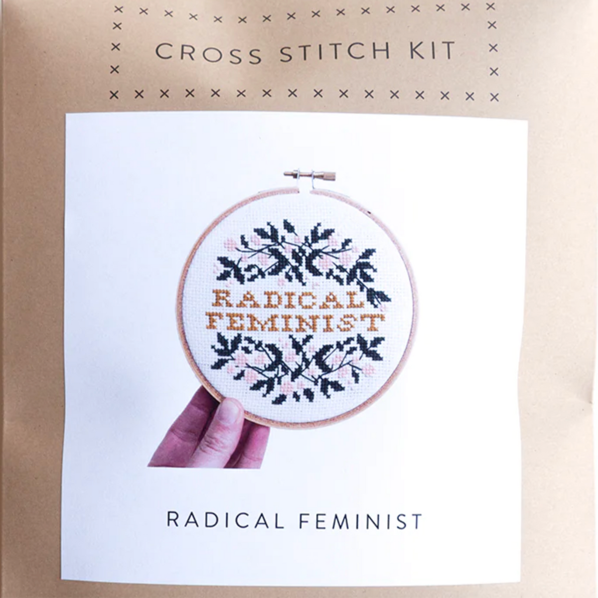 The packaging for the Radical Feminist Cross Stitch Kit by Junebug & Darlin. It is a brown kraft paper envelope with a picture of the completed kit on the front. It says Radical Feminist in the middle and is surrounded by pretty floral vines. 