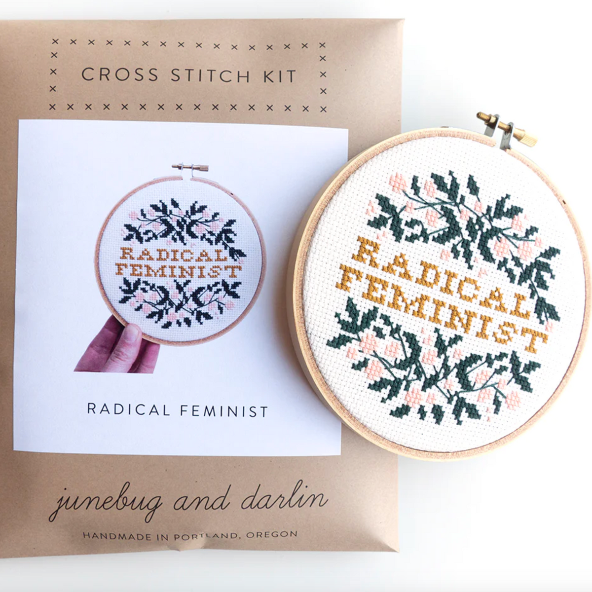 A stitched up example of the Radical Feminist Cross Stitch kit by Junebug & Darlin. It is sitting on top of the package for the kit which is a kraft brown paper envelope with a photo of the completed kit on the front. 