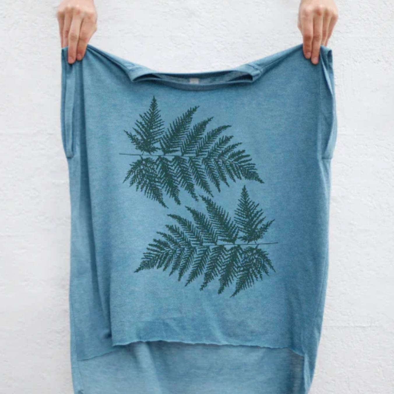 Blue rolled cuff top with fern leaf design held by a person against a white brick wall.