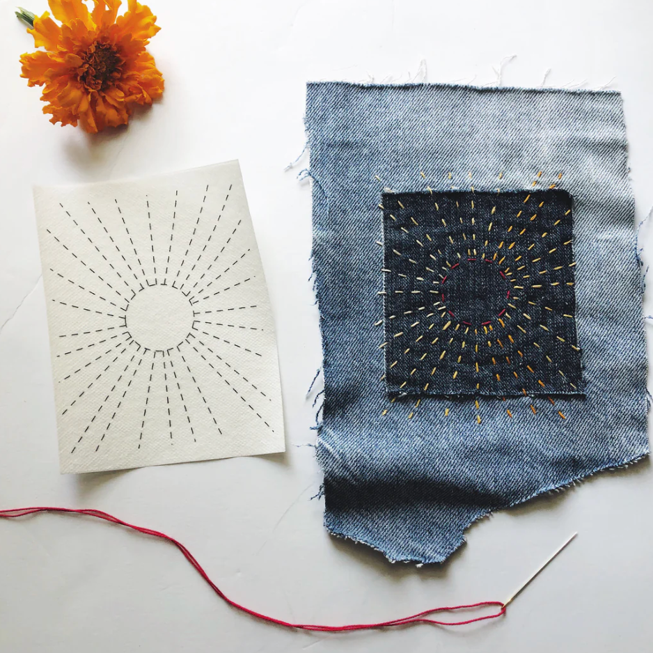 A design that looks like a sun is stitched onto denim. A flower and a threaded needle are on the table as well as the pattern sheet.