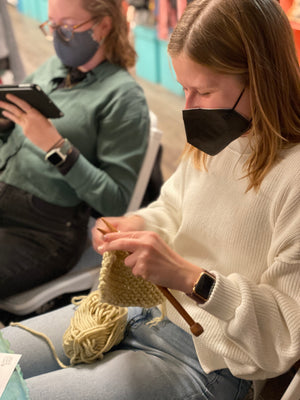 Woman knitting with a mask on, sitting next to another person using a phone.