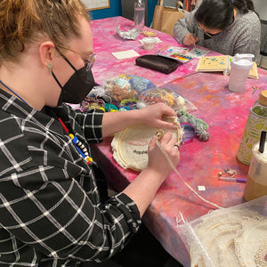Person working on a craft project at a table with various materials and another person in the background.