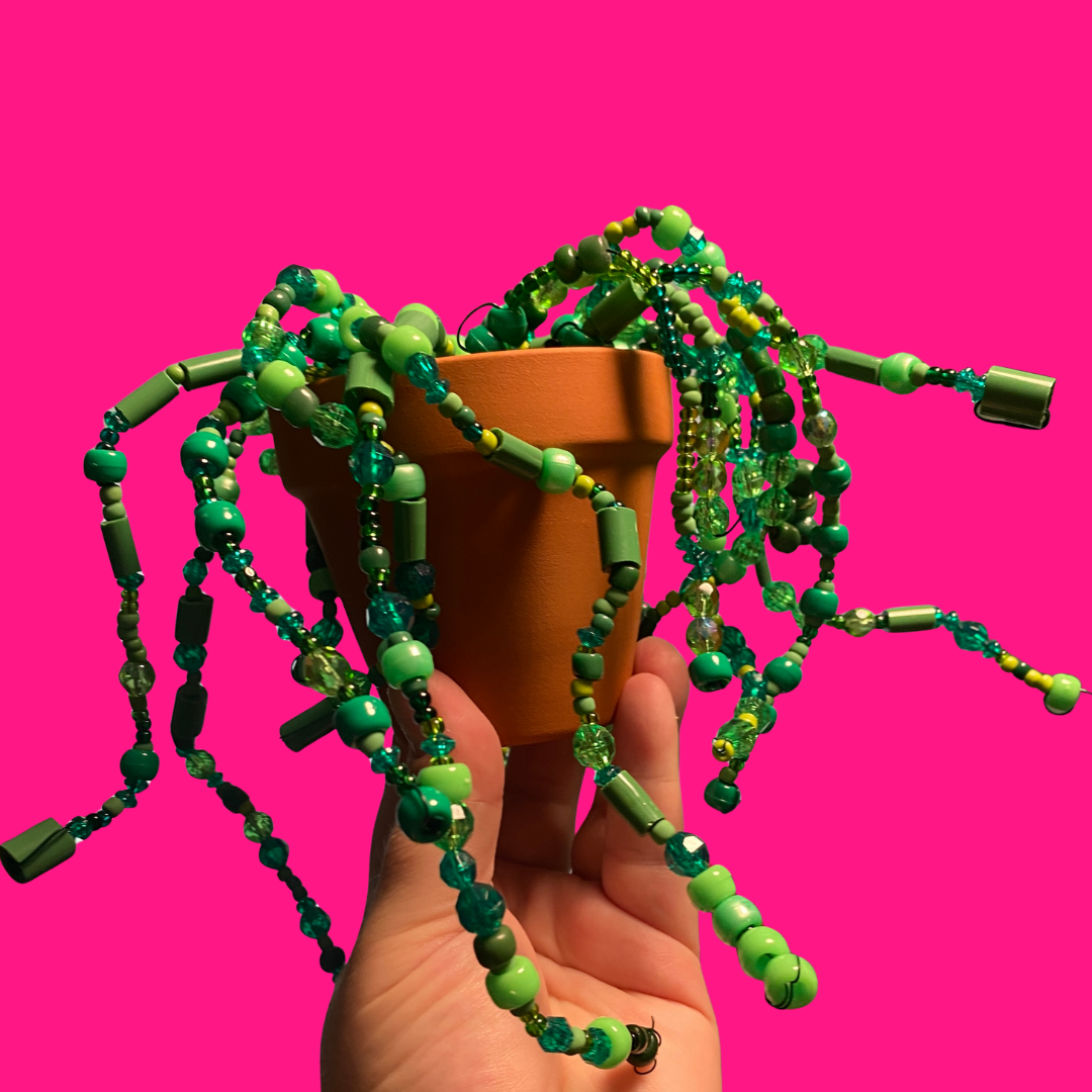 A hand holding up a small terracotta pot with a beaded plant inside. There is a hot pink background. 