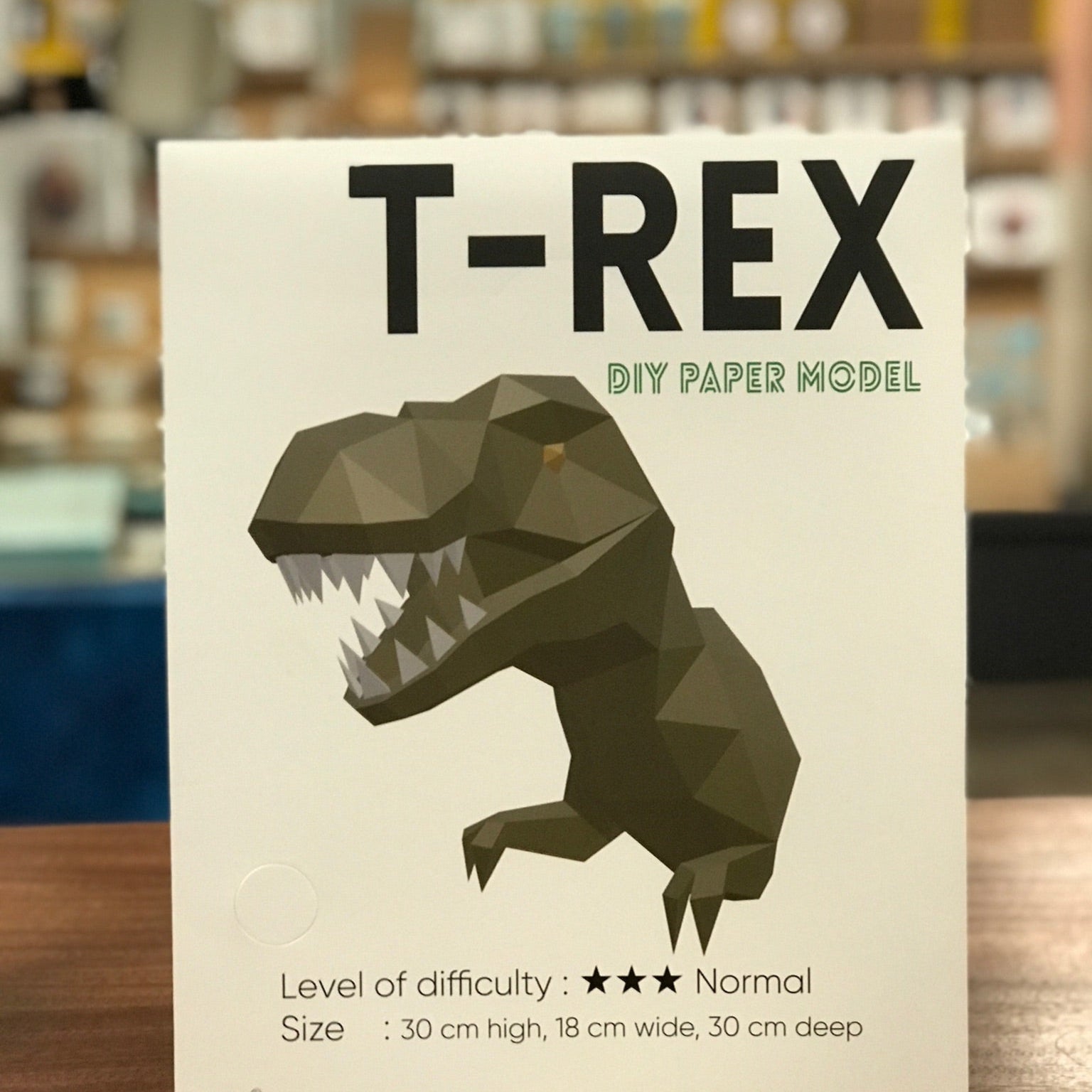 Packaging for T-Red Paper Craft Kit. It is white and has T-Rex written in bold black capitol letters. It has an example of the completed dinosaur paper craft.