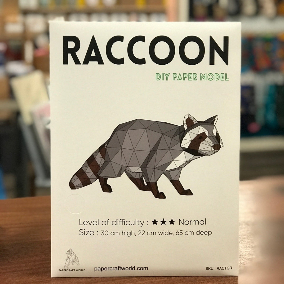 Packaging for the Raccoon paper craft. It is white with "Raccoon" written is bold capitol letters in black. It has an illustration of a completed raccoon paper craft. 